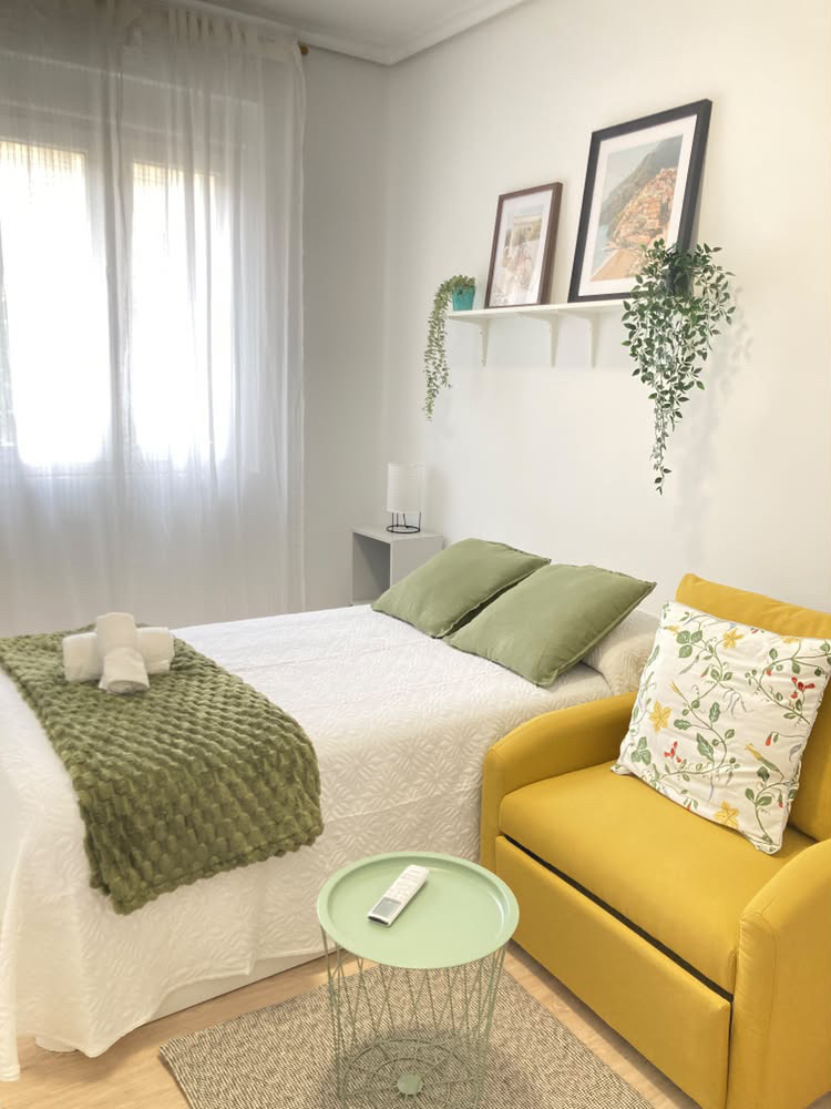 Habitación D: cama con manta verde, sillón mostaza y láminas decorativas